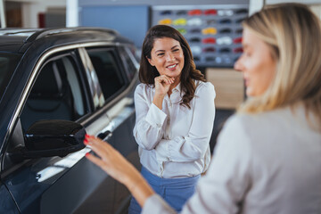 Businesswoman looking for a new car, standing next to a new SUV indoors at a car dealer. Beautiful young caucasian female client customer choosing new car, trying checking its options