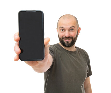 Portrait oh happy man with beard wearing brown t-shirt shows something interesting on the smartphone screen, isolated on white background. Bald guy holding cell phone smiling and posing in studio.