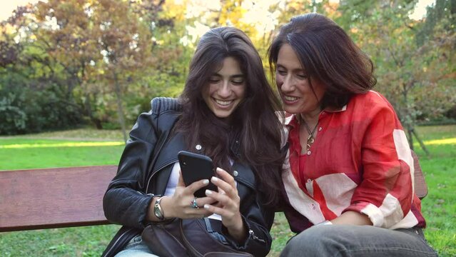 Happy young woman and adult woman watching photos on a mobile phone sitting on a bench