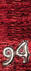 Silver foil balloon number 94 on a background of red tinsel decoration. Birthday greeting card,...