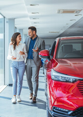  Young family selecting vehicle, looking at modern automobile at showroom. Car sales business. Beautiful young couple at car showroom choosing a new car to buy.