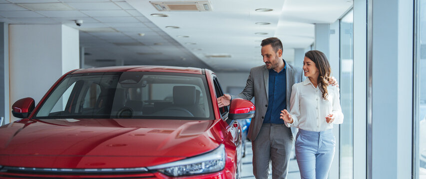 Portrait of a beautiful young couple choosing a new car at a car dealership. Buying a long-awaited car. Couple buying the car. Pair in a car salon. Husband with vife choose the automobile