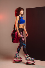 Curly woman in sportswear and bag wearing kangoo jumpers posing in studio on background - 718041260