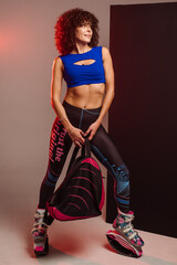 Curly woman in sportswear and bag wearing kangoo jumpers posing in studio on background - 718041439