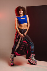 Curly woman in sportswear and bag wearing kangoo jumpers posing in studio on background - 718041441
