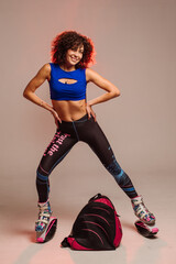 Curly woman in sportswear and bag wearing kangoo jumpers posing in studio on background - 718041456