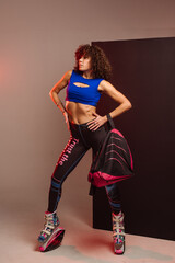 Curly woman in sportswear and bag wearing kangoo jumpers posing in studio on background - 718041490