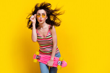 Portrait of excited girl fluttering hair touch glasses hold skateboard look at offer empty space isolated on yellow color background