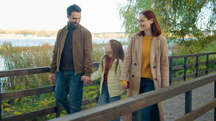 Happy Caucasian family parents couple with adopted custody adorable daughter child girl kid weekend in park autumn outdoors near lake river bridge walking talking smiling together walk holding hands