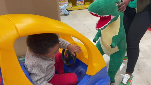 Back child, 3y.o, playing with his mother with a huge plush dinosaur inside a toy store
. He is seated inside a big plastic toy car. 4K