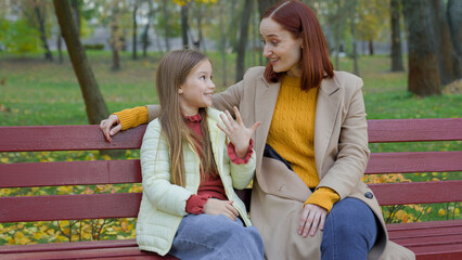 Fototapeta premium Caucasian woman mother mom talking with child girl kid daughter teen schoolgirl family talk friendly conversation motherhood childhood childcare sitting at city park autumn at bench laugh smile news