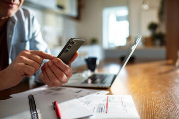 Middle aged man using smartphone at home office with laptop