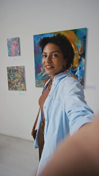 Medium vertical selfie shot of young African American woman shooting video on smartphone for social media while visiting contemporary art exhibition in museum, and showing paintings to followers