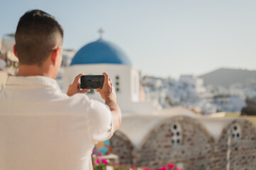Tourist taking photo on smartphone of Oia cityscape on vacation - obrazy, fototapety, plakaty
