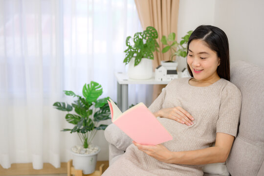 Asian pregnancy woman relaxes and reads a book on the sofa in the living room at home. Carrying a large belly Happy pregnant woman at her home before giving birth