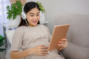 Asian pregnant woman using tablet Listen to music and relax on the sofa in the living room at home. Carrying a large belly Happy pregnant woman at her home before giving birth