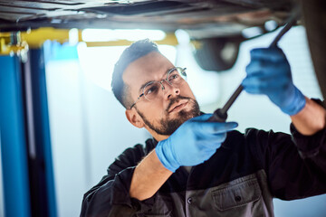 Focused mechanic man, checking something underneath the car, wearing protective gloves.