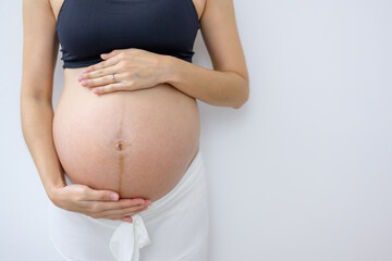 Close-up photo of pregnant woman with pregnancy holding her large belly standing against the wall in a white room at home.