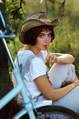 Young beautiful woman with short hair in cowboy hat and blue overall sitting near tree next to bicycle. Romantic portrait of village girl holding her finger near lips