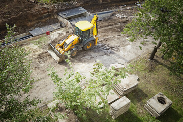 An excavator in the middle of a construction site with green trees around. Repair of urban communications, work at the construction site