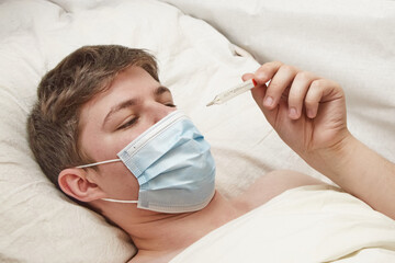 A young man wearing a medical mask is lying in bed under a blanket and looking at a thermometer...