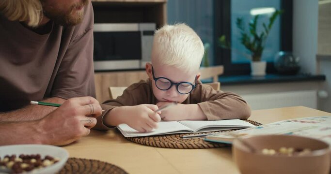 Close-up shot of a little albino boy with white hair wearing blue glasses puts down his pen and raises his two palms up to count on his fingers during his math homework