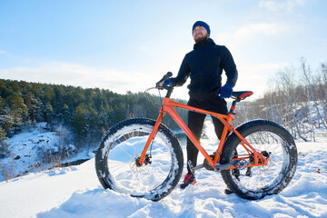 Positive bearded young cyclist holding fatbike and standing on mountain reflecting about life