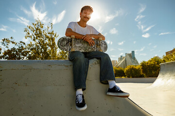 Smiling young male skateboarder holding skateboard sitting on ramp in skate park 