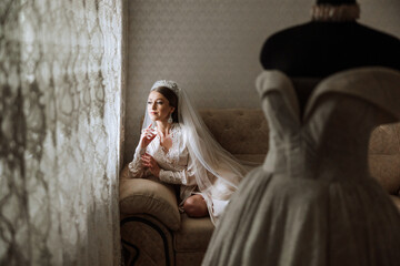 A beautiful bride with a long veil in her room, wearing a robe. Wedding dress on a mannequin. The...
