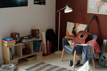 New acoustic guitar standing on armchair with pile of clothes in front of record player surrounded...