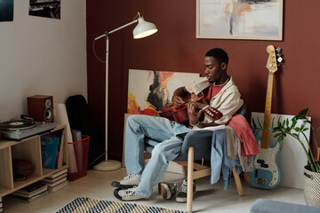 Young African American male musician in casualwear sitting in comfortable armchair in the corner of...