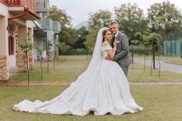 Fototapeta premium A handsome groom embraces his bride in a lush white dress and smiles in a beautiful outdoor setting. Under the open sky. High quality photo. A newlywed couple poses together on a sunny summer day.