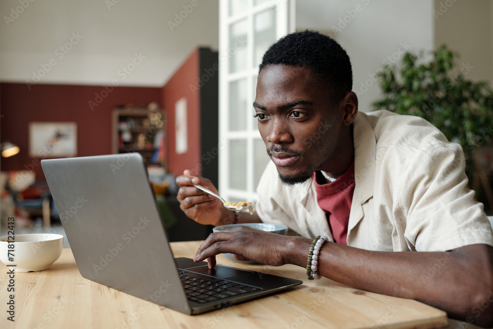 Poster serious young man sitting by table in front of laptop screen while having breakfast and watching onl