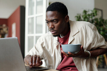 Serious African American businessman with bowl of cornflakes in hand looking at laptop screen while...
