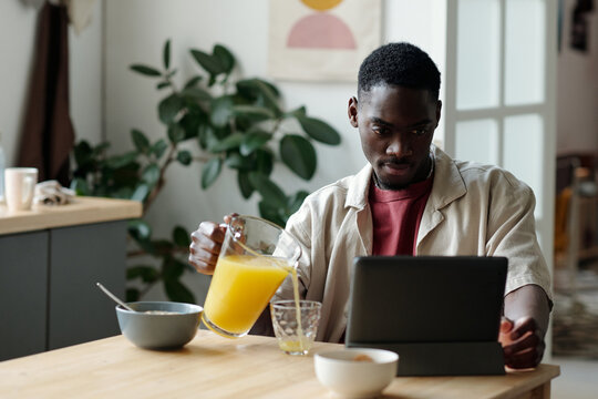 African American businessman or student looking at tablet screen while pouring fresh orange juice into glass during breakfast