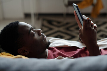 African American man with smartphone lying on couch in front of camera and watching online video or...