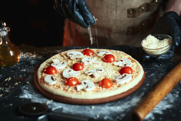 The chef sprinkles cheese on a pizza with mushrooms and tomatoes. Pizzaiolo in a leather apron prepares pizza.