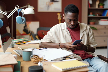 Young male freelancer looking at smartphone screen while sitting by workplace with papers and notepads and organizing work
