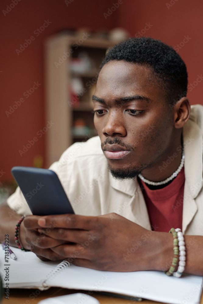 Poster young serious male student looking at smartphone screen while sitting by table with open copybook in