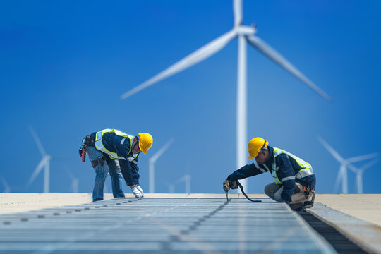 Worker Technicians are working to construct solar panels system on roof with wind turbine on background. Installing solar photovoltaic panel system. Renewable clean energy technology concept.