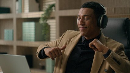 Relaxed carefree African American man guy entrepreneur male businessman listen music on laptop in headphones relax take break office work pause dancing enjoy song melody smiling funny dance moving fun