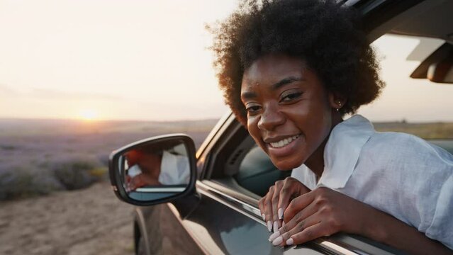 Black young woman afro hair car trip relax hand plays rejoices with rays of sun sitting in car looks in side mirror at camera in lavender field at sunset in summer on vacation. Go Everywhere freedom