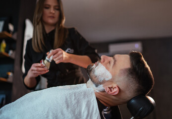 Young longhaired female Barber gently applies shaving cream foam on bearded man's face bristle...