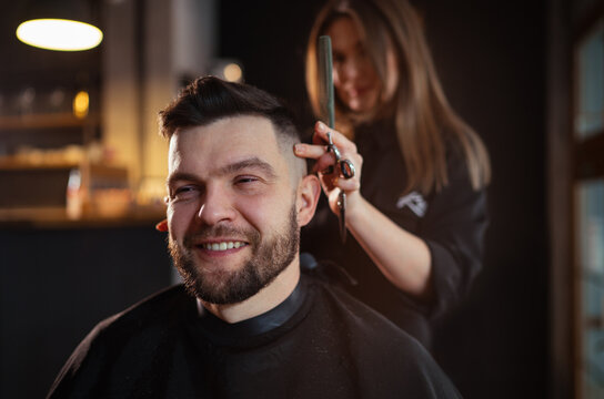 Smiling bearded Man portrait while undercut hairstyle hairdressing in Hair Salon by young hairdresser female. Modern low light black style barber shop interior. Haircare service small business concept