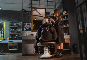 Beautiful Young female barber making men's hairstyle using Haircut Clipper in Modern loft style barber shop interior. Haircare services local small businesses, cosmetics and personal care industry. - obrazy, fototapety, plakaty