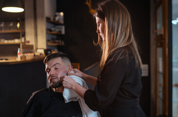 Long-haired young female Barber gently shaving bearded man face bristle while he in chair. She removing cream foam remains from cheeks.Hair and skin care service, local small business concept.