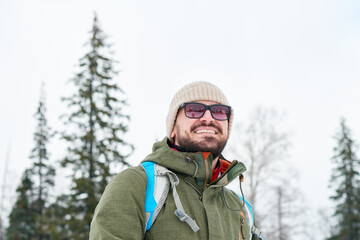 Fototapeta na wymiar Cheerful handsome bearded backpacker in sunglasses looking into distance enjoying beauty of winter landscape