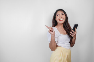 A cheerful Asian woman is holding her smartphone and pointing copy space beside her, isolated by white background.