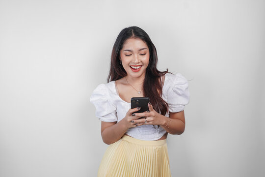 A cheerful Asian woman is smiling and holding her smartphone, isolated by white background.