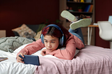 Teenage girl in headphones looking at screen of smartphone while relaxing on bed on weekend and...